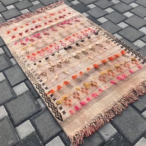 May include: A rectangular, beige rug with a striped pattern and colourful accents. The rug features rows of orange, pink, and red tufts, with black geometric designs on the border. The rug has a fringed edge.