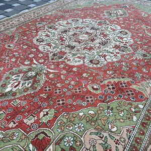 May include: A large, rectangular, vintage rug with an intricate floral pattern. The rug features a central medallion design in shades of red, green, and cream. The border is a lighter beige with a repeating floral motif. The rug is placed on a dark gray surface.