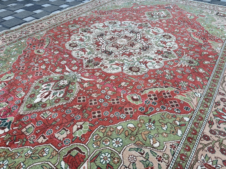 May include: A large, rectangular, vintage rug with an intricate floral pattern. The rug features a central medallion design in shades of red, green, and cream. The border is a lighter beige with a repeating floral motif. The rug is placed on a dark gray surface.
