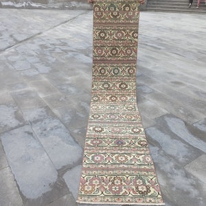 Pode incluir: Um tapete longo e estreito com fundo creme e padrões florais e geométricos intrincados em tons de rosa, verde e marrom. O tapete está dobrado ao meio e está sobre um piso de concreto cinza.