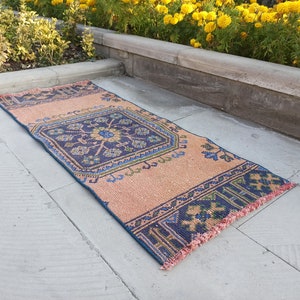 May include: A peach-colored rug with a blue and green geometric pattern. The rug has a fringe border and is lying on a gray concrete surface.