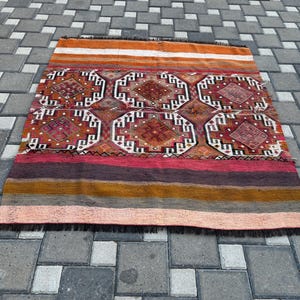 May include: A rectangular woven rug featuring a central section with six repeating octagonal geometric motifs in shades of red, brown, and white. The top and bottom borders consist of horizontal stripes in orange, pink, purple, and yellow.