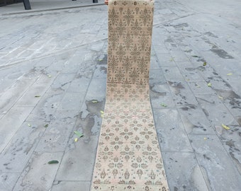 May include: A long, narrow, cream-coloured rug with a repeating pattern of brown and green floral designs. The rug is folded in half and lying on a grey concrete surface.