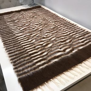 Puede incluir: Una alfombra de pelo sintético de rayas marrones y blancas con flecos. La alfombra tiene una textura suave y esponjosa y es perfecta para añadir un toque de calidez y estilo a cualquier habitación.