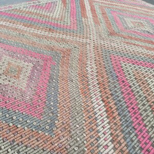 May include: A close-up of a woven rug with a geometric pattern in shades of pink, gray, and beige. The rug has a diamond shape in the center and is made of a soft, textured material.