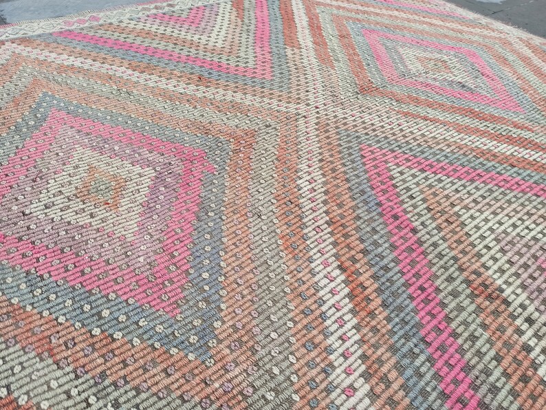 May include: A close-up of a woven rug with a geometric pattern in shades of pink, gray, and beige. The rug has a diamond shape in the center and is made of a soft, textured material.