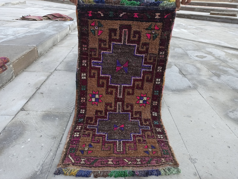 May include: A vintage Turkish rug with a geometric pattern in shades of brown, red, pink, blue, and green. The rug has a fringed border and is folded in half.