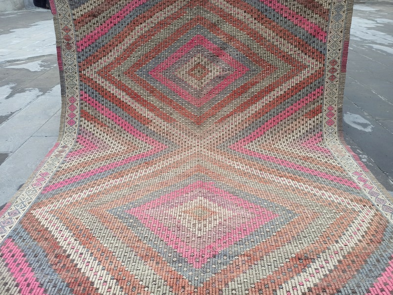 May include: A pink, gray, and brown patterned rug with a diamond shape in the center. The rug is folded in half and the edges are visible.