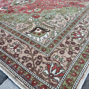 May include: Close-up of a section of a vintage rug with a detailed floral pattern. The rug features a central field of red and green, framed by a beige border with floral and geometric designs. The rug's edges show a fringe.