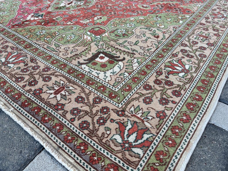 May include: Close-up of a section of a vintage rug with a detailed floral pattern. The rug features a central field of red and green, framed by a beige border with floral and geometric designs. The rug's edges show a fringe.