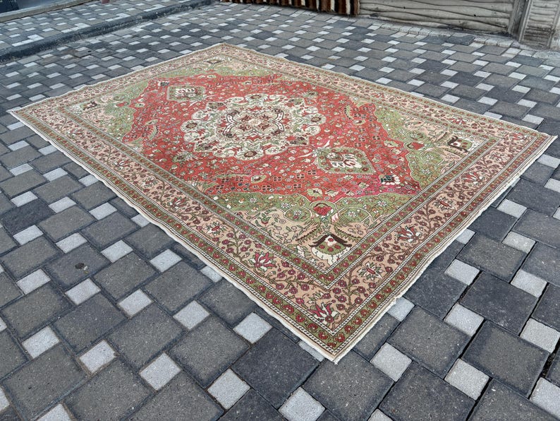 May include: A rectangular, vintage rug with a central medallion design. The rug features a red field with green and cream accents, and a detailed border. The rug is placed on a gray tiled surface.