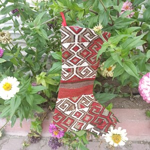 Puede incluir: Una media de Navidad roja, blanca y marrón con un diseño geométrico. La media está colgada en un jardín con plantas verdes y flores.