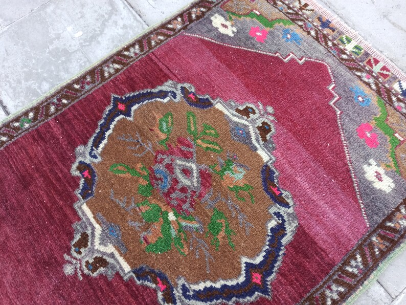 Puede incluir: Una alfombra turca vintage con fondo rojo y un medall&oacute;n central con un dise&ntilde;o floral en tonos marrones, verdes y azules. La alfombra tiene un borde con un patr&oacute;n geom&eacute;trico en tonos grises y blancos.