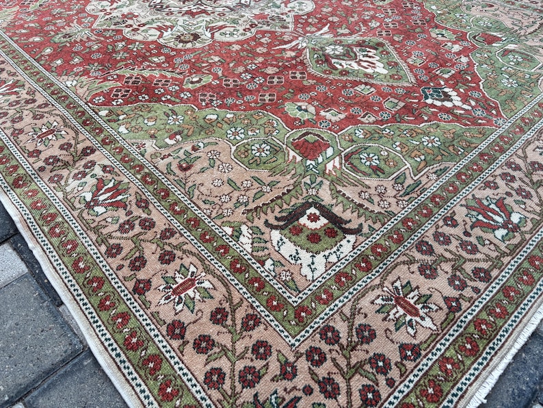 May include: Close-up of a section of a vintage rug with a detailed floral pattern. The rug features a red central field with green and beige accents. The border is a mix of beige, green, and red floral designs.