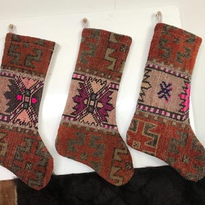 May include: Three handmade Christmas stockings with a rustic, vintage aesthetic. The stockings feature a woven design in shades of rust, brown, and pink, with geometric patterns and a central floral motif. Each stocking is approximately 18 inches long.