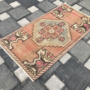 Può includere: Un tappeto rettangolare in stile vintage con un design geometrico centrale nei toni del pesca, marrone e azzurro. Il tappeto presenta motivi intricati e un bordo sfrangiato. Il tappeto è posizionato su una superficie piastrellata grigia e bianca.