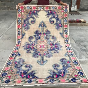 May include: A vintage rug with a cream background and a colorful floral and geometric pattern in shades of pink, blue, and green. The rug has a fringed edge.