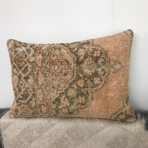 May include: Rectangular decorative pillow with a vintage, distressed look. The pillow features a patterned design in shades of brown, green, and peach. The pillow is placed on a light-colored, textured surface.