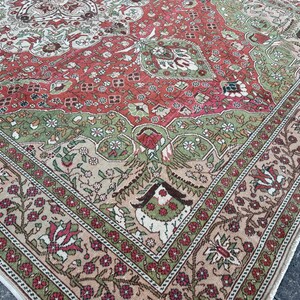 May include: A detailed shot of a large, rectangular rug with an intricate floral design. The rug features a color palette of red, green, and beige, with a prominent border. The pattern includes floral and geometric elements.