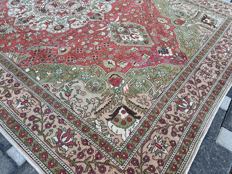 May include: A detailed shot of a large, rectangular rug with an intricate floral design. The rug features a color palette of red, green, and beige, with a prominent border. The pattern includes floral and geometric elements.
