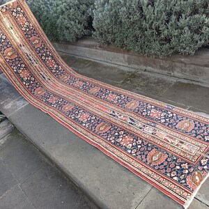 Puede incluir: Una alfombra larga y estrecha con un diseño persa tradicional. La alfombra tiene un fondo azul oscuro con patrones florales rojos y crema. La alfombra está colocada sobre un conjunto de escalones de piedra.