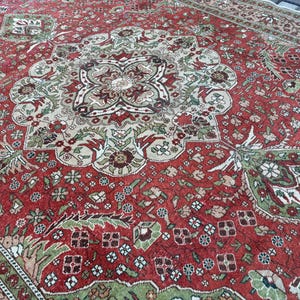 May include: A detailed, antique-style rug with a rich red background and intricate floral and geometric patterns in shades of green, beige, and brown. The rug features a central medallion design and a border with repeating motifs, showcasing a classic, ornate aesthetic.