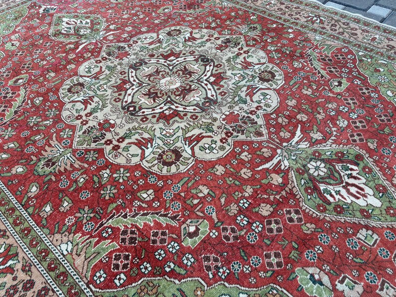 May include: A detailed, antique-style rug with a rich red background and intricate floral and geometric patterns in shades of green, beige, and brown. The rug features a central medallion design and a border with repeating motifs, showcasing a classic, ornate aesthetic.