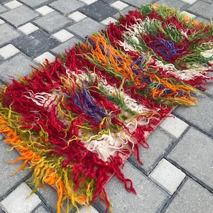 Könnte beinhalten: Ein rechteckiger, zotteliger Teppich mit einer lebendigen Farbmischung. Der Teppich hat lange, strukturierte Fasern in Rot-, Orange-, Gelb-, Grün-, Weiß- und Lilatönen. Der Teppich liegt auf einer grauen, steinartigen Oberfläche.