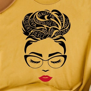 May include: Mustard yellow t-shirt featuring a black graphic of a woman's face with glasses and red lipstick. The woman's hair is styled in a bun with words like "fierce" and "beautiful" incorporated into the design. A brown hat rests on the shirt.