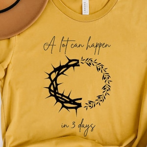 May include: Mustard yellow t-shirt with the text "A lot can happen in 3 days" and a black graphic of a crown of thorns and a wreath. A brown hat and a white mug are also in the image.