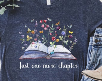 May include: A navy blue t-shirt with a graphic of an open book with flowers and butterflies growing from it. The text "Just one more chapter" is printed below the book. The shirt is laid out on a white surface with a pair of light brown ankle boots and blue jeans.