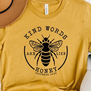 May include: Mustard yellow t-shirt with a black graphic of a bee and the text "KIND WORDS ARE LIKE HONEY PROVERBS 16:24". A brown hat with a black band is on top of the shirt.