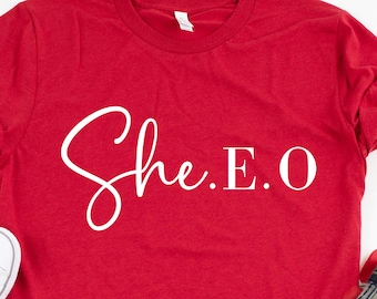 Puede incluir: Camiseta roja con texto blanco que dice "She.E.O". La camiseta está sobre una superficie de madera blanca con pétalos de rosa roja esparcidos alrededor. También hay un par de zapatillas blancas y un par de pantalones vaqueros azules en la imagen.