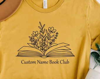 May include: A mustard yellow t-shirt with a black outline of an open book with flowers growing out of it. The text "Custom Name Book Club" is printed below the book.