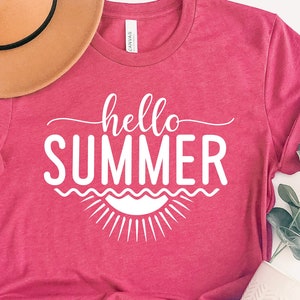 May include: A pink t-shirt with the text "hello summer" in white lettering. The shirt is styled with a brown hat and a sprig of greenery.