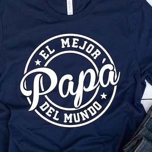 May include: A navy blue t-shirt with a white circle graphic that says "El Mejor Papa Del Mundo" which translates to "The Best Dad in the World".