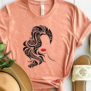 May include: A peach-colored t-shirt with a black silhouette of a woman's head with the words "fuerte", "bella", "poderosa", "trabajadora", "mujer", "latina", "guerrera", "unica", "hermosa", "alma" written around her hair.