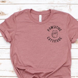 Puede incluir: Una camiseta de color malva con un gr&aacute;fico negro de un gato con el texto "Pawsitive Cattitude" en la parte delantera.