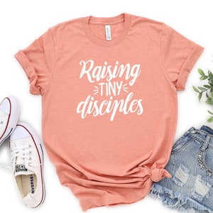 May include: A peach colored t-shirt with the text "Raising Tiny Disciples" in white lettering.