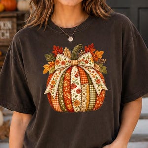 May include: A dark brown short-sleeve t-shirt featuring a central graphic of a pumpkin decorated with a large bow and autumn leaves. The pumpkin design incorporates various patchwork patterns including floral, plaid, and solid orange, red, and green colors.