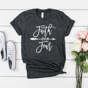 May include: A dark gray t-shirt with the text "Faith over Fear" in white lettering with a white arrow and feather design. The shirt is on a white wooden background with white sneakers and pink flowers.