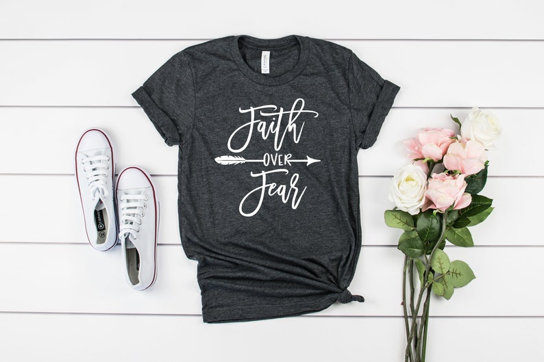May include: A dark gray t-shirt with the text "Faith over Fear" in white lettering with a white arrow and feather design. The shirt is on a white wooden background with white sneakers and pink flowers.