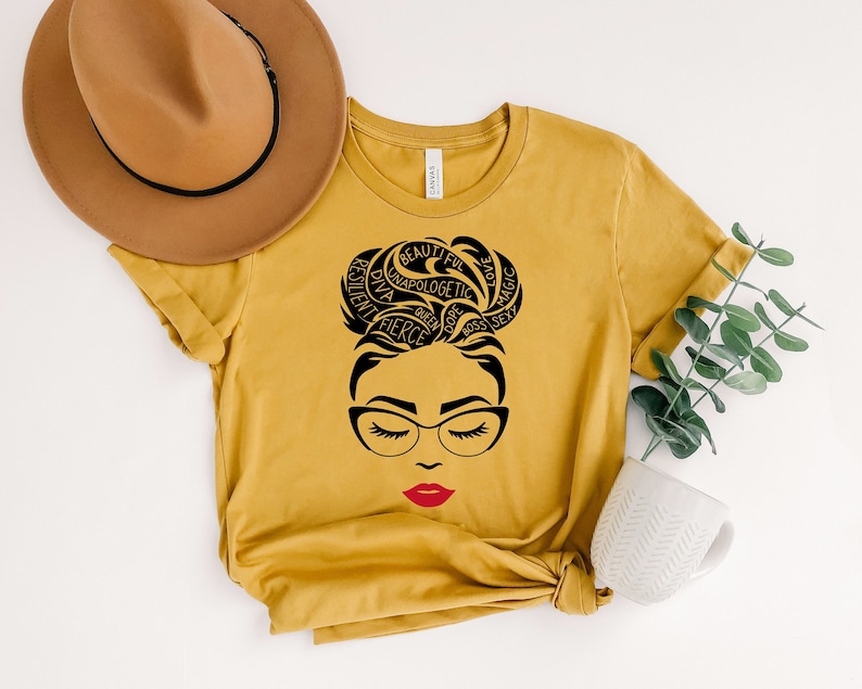 May include: Mustard yellow t-shirt featuring a black graphic of a woman's face with glasses and red lipstick. The woman's hair is styled in a bun with words like "fierce" and "beautiful" incorporated into the design. A brown hat rests on the shirt.