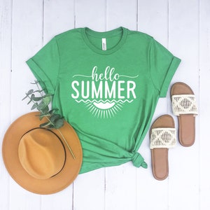 May include: A green t-shirt with the text "hello summer" in white lettering. The shirt has a white sun graphic with waves underneath the text. The shirt is laid flat on a white wooden surface with a brown hat and two pairs of sandals.