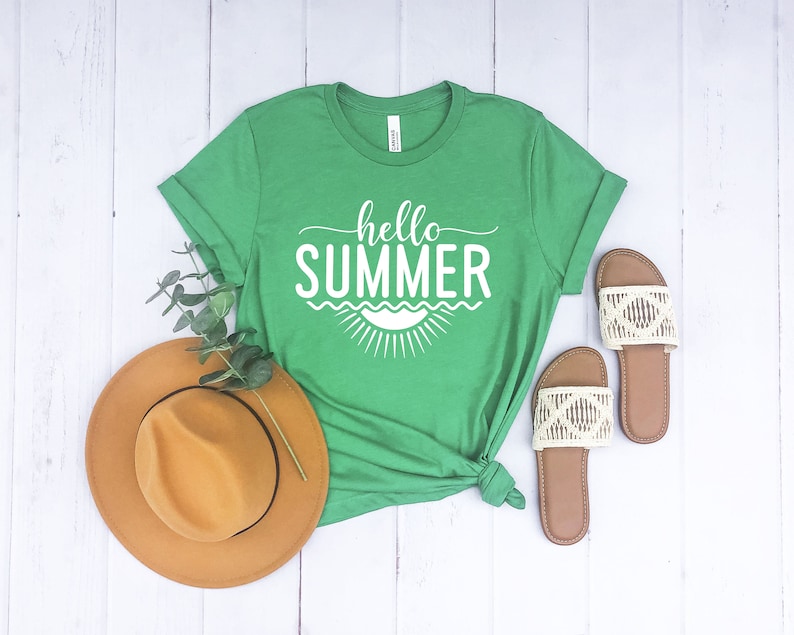 May include: A green t-shirt with the text "hello summer" in white lettering. The shirt has a white sun graphic with waves underneath the text. The shirt is laid flat on a white wooden surface with a brown hat and two pairs of sandals.