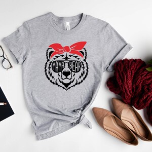 以下が含まれることがあります： サングラスと赤いバンダナをつけたクマのグラフィックが特徴のグレーのTシャツで、「MAMA BEAR」の文字が入っています。メガネ、ノート、靴など、アクセサリーも一緒にコーディネートされています。