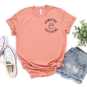 Puede incluir: Una camiseta de color melocot&oacute;n con un gr&aacute;fico negro de un gato con el texto "Pawsitive Cattitude". La camiseta est&aacute; colocada sobre un fondo blanco con un par de zapatillas blancas y pantalones cortos de mezclilla.