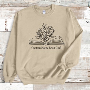 May include: A sand-colored sweatshirt with a black outline of an open book with flowers growing out of it. The text "Custom Name Book Club" is printed below the book.