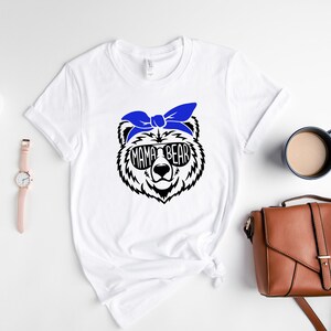 以下が含まれることがあります： サングラスと青いヘアバンドをつけた白黒のクマのグラフィックが特徴の白いTシャツ。「MAMA BEAR」の文字がサングラスにプリントされています。シャツは、アクセサリーと一緒に白い表面に表示されています。