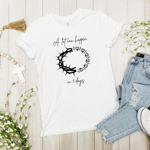 May include: White t-shirt featuring the text "A lot can happen in 3 days" and a black graphic of a crown of thorns and a wreath. The shirt is styled with blue jeans and white trainers.